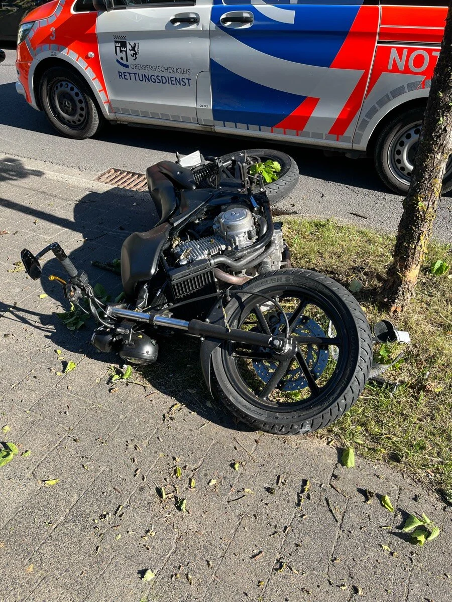 Ein schwarzes Motorrad liegt umgestürzt auf einem Gehweg neben einem Rettungsfahrzeug des Oberbergischen Kreises bei sonnigem Wetter.