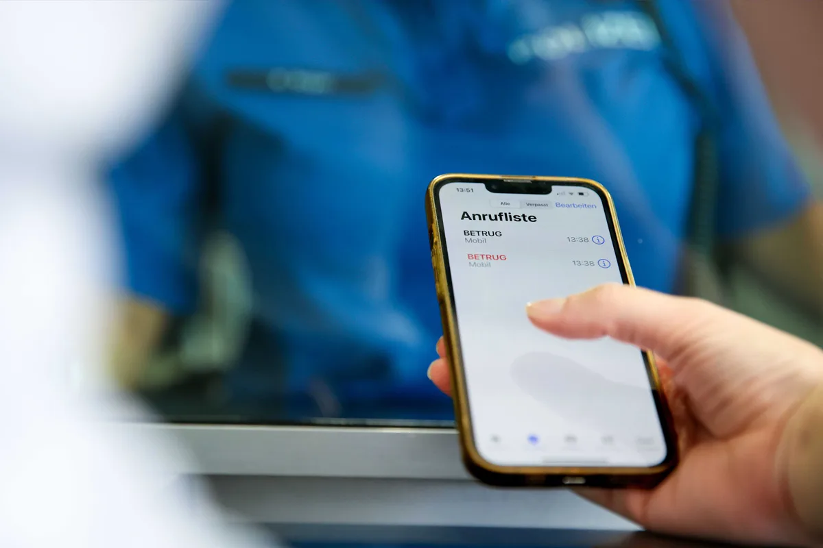 Eine Person hält ein Smartphone in der Hand, auf dem zwei Einträge mit 'Betrug' in der Anrufliste angezeigt werden. Im Hintergrund ist ein Polizist teilweise erkennbar.