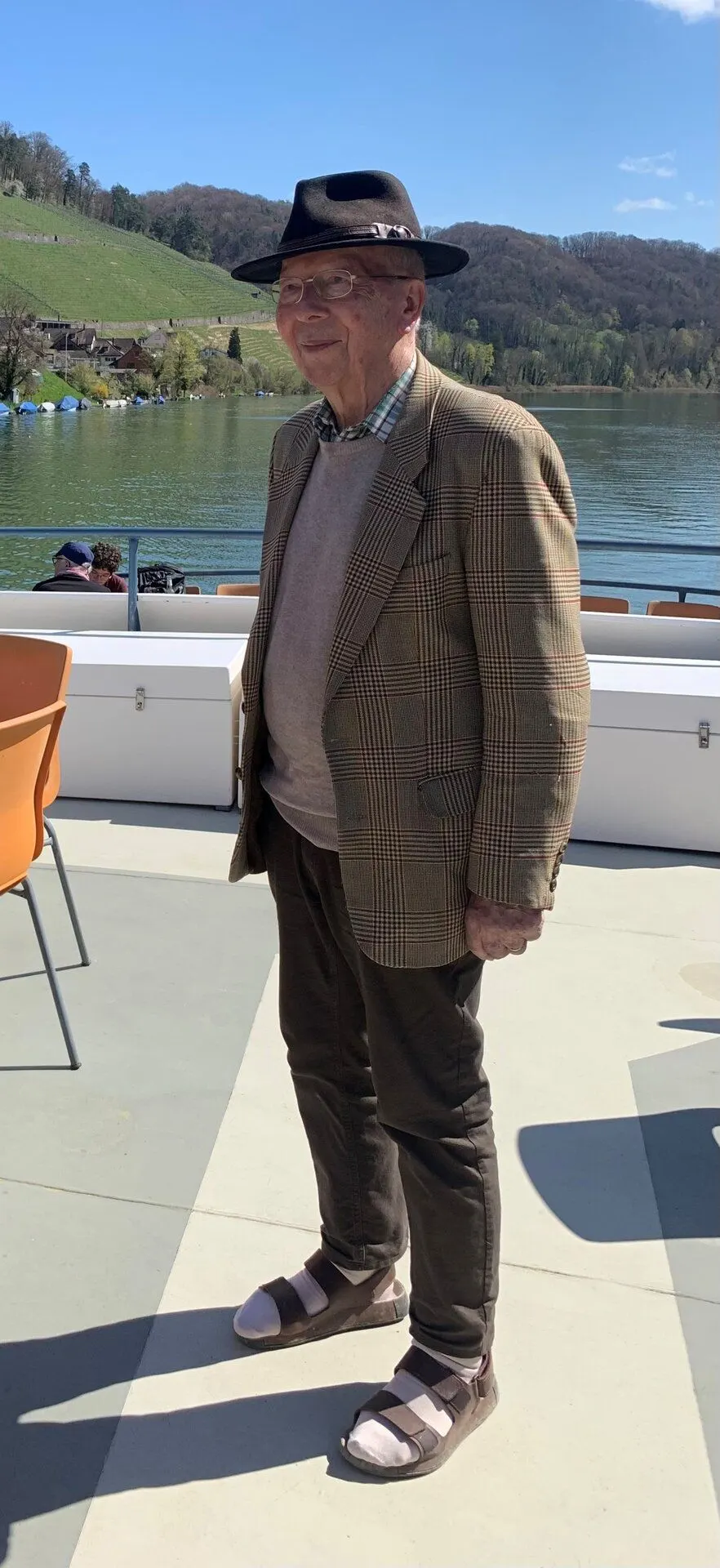 Ein älterer Mann mit Hut und Jackett steht auf einem Bootssteg am Fluss. Im Hintergrund sind Hügel und Bäume zu sehen.
