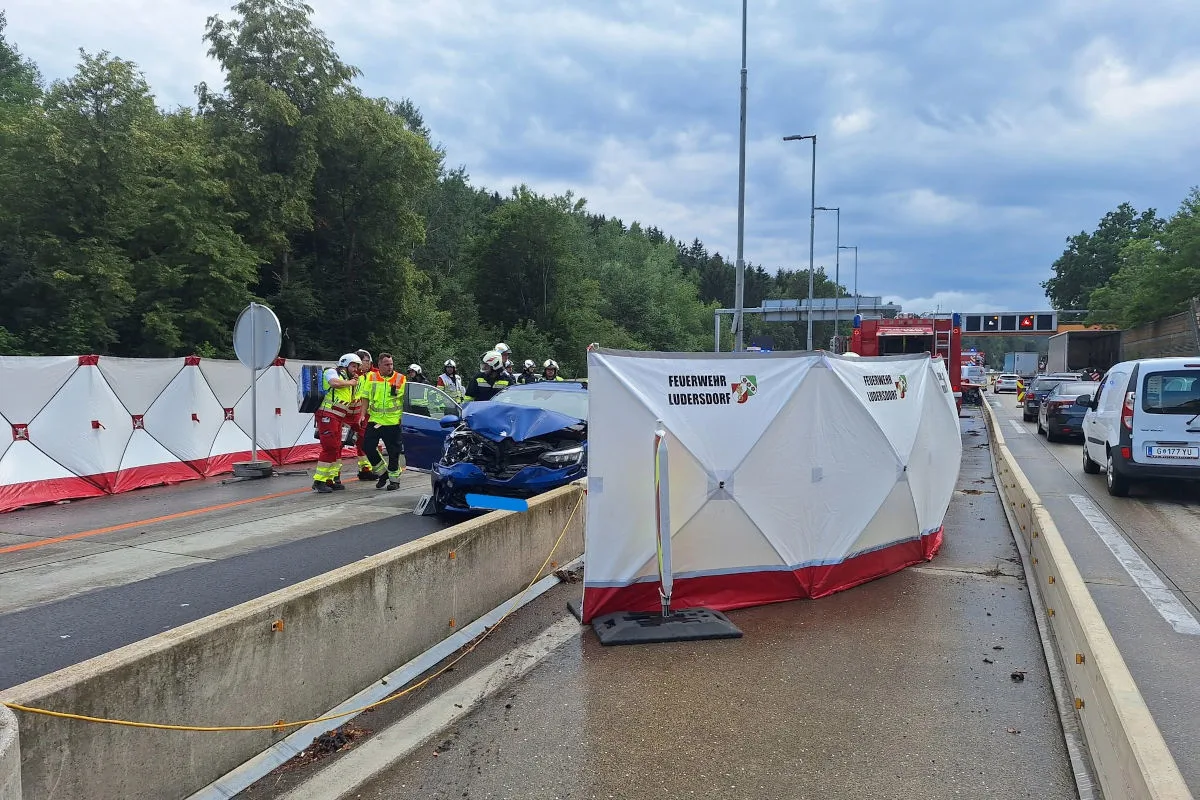 Ein blaues Auto mit Frontschaden steht auf einer Autobahn, umgeben von Rettungskräften und Absperrungen. Eine mobile Sichtschutzwand und Feuerwehrleute sind ebenfalls zu sehen.