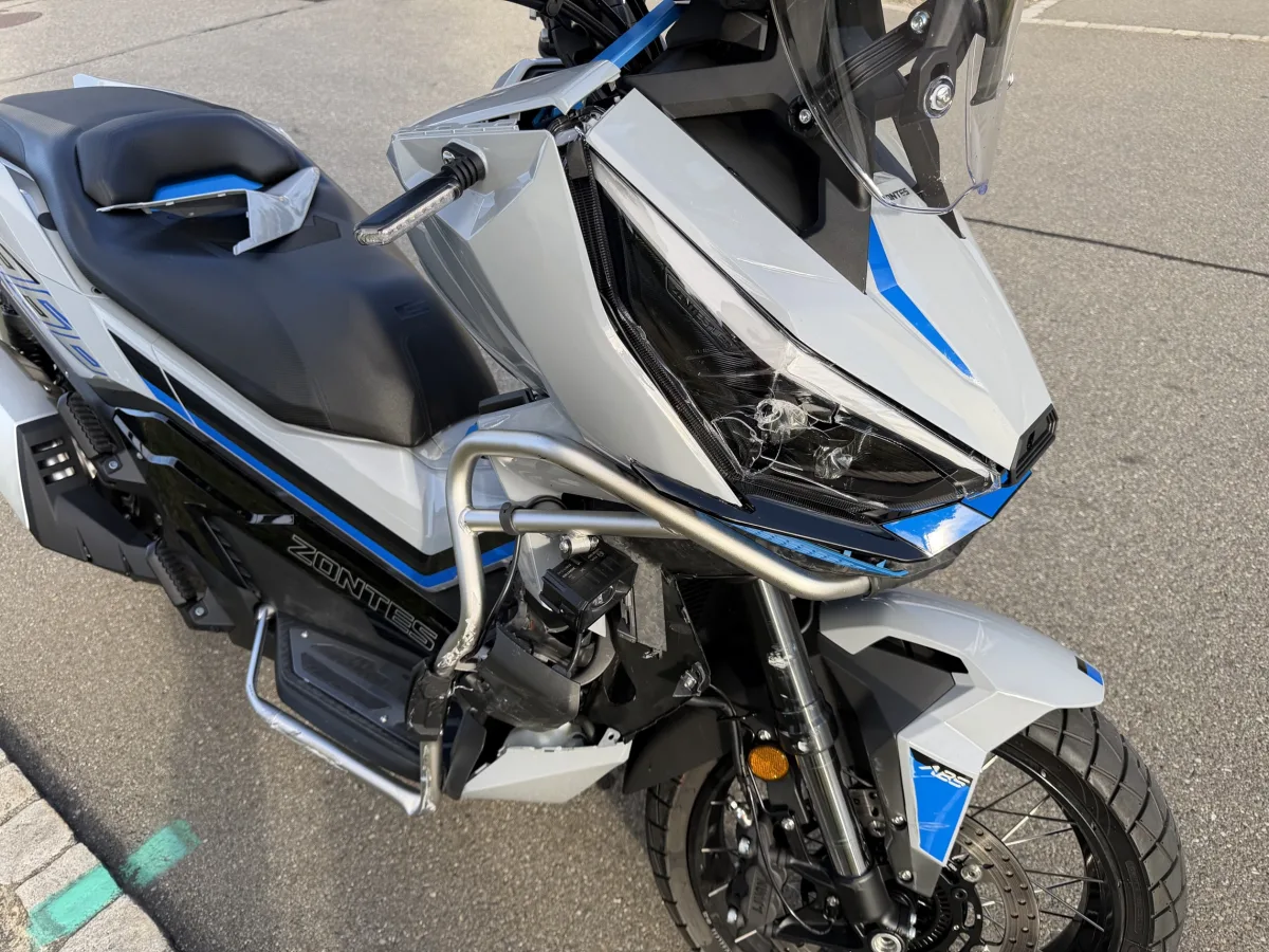 Ein graues Motorrad, Marke Zontes, zeigt einen gebrochenen Scheinwerfer auf einer Straße. Der Lack ist grau mit blauen Akzenten.