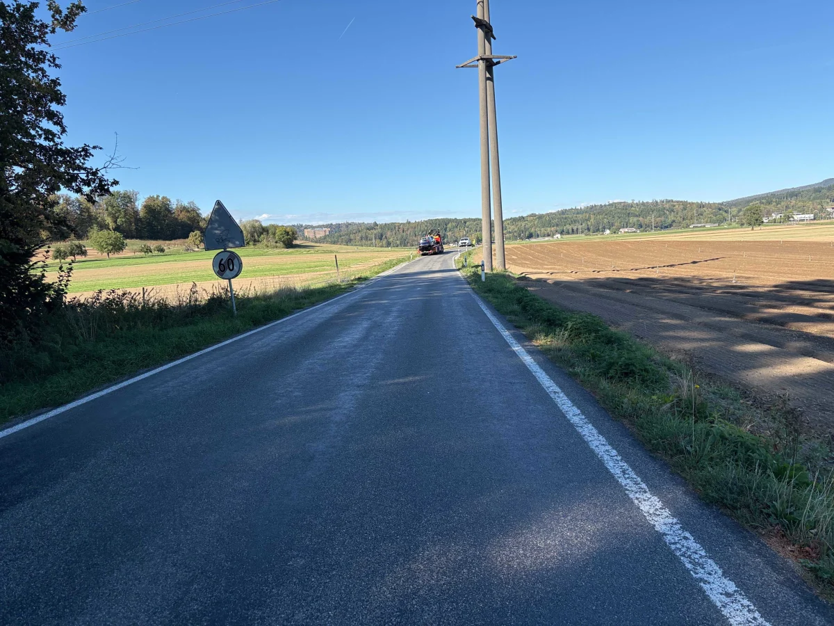 Eine Landstraße verläuft zwischen Feldern, ein Traktor ist in der Ferne sichtbar unter klarem Himmel.