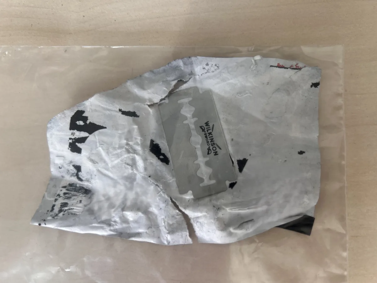 Eine einzelne Rasierklinge liegt auf einem zerknitterten weißen Stück Papier, verpackt in einer klaren Tüte.