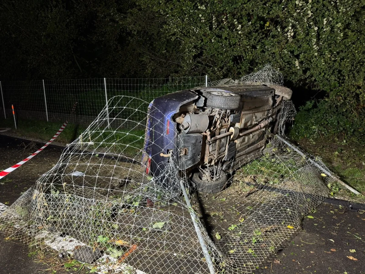 Ein blaues Auto liegt seitlich in einer Böschung, umgeben von beschädigtem Zaun und Vegetation. Die Szene ist nachts aufgenommen.