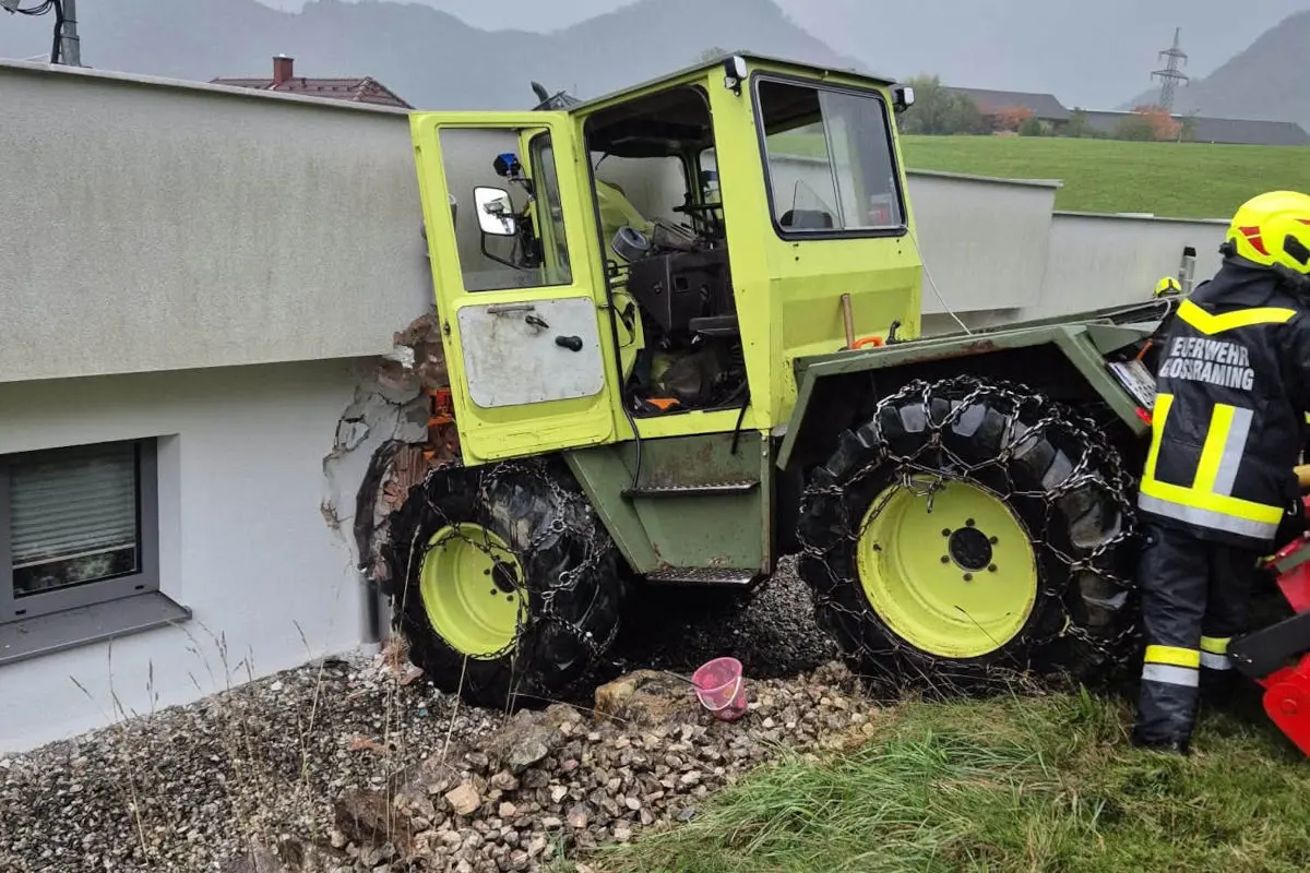 Ein grüner Traktor steckt in einer Gebäude-Wand, und ein Feuerwehrmann steht daneben. Der Traktor hat Ketten auf den Reifen.