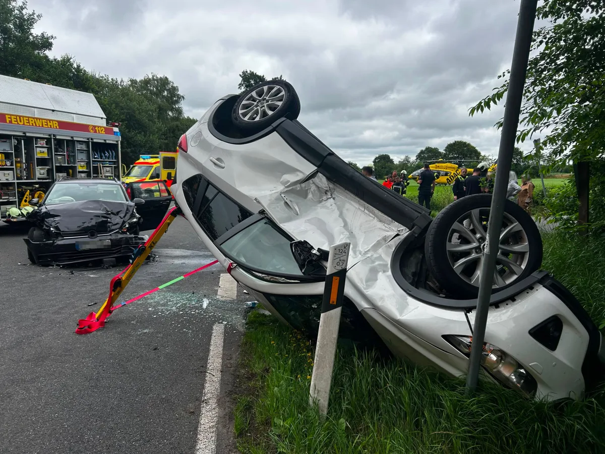 Ein weißes Auto liegt kopfüber neben der Straße, während ein weiteres Fahrzeug mit Frontschaden sichtbar ist. Feuerwehrfahrzeug und Einsatzkräfte sichern die Unfallstelle ab.
