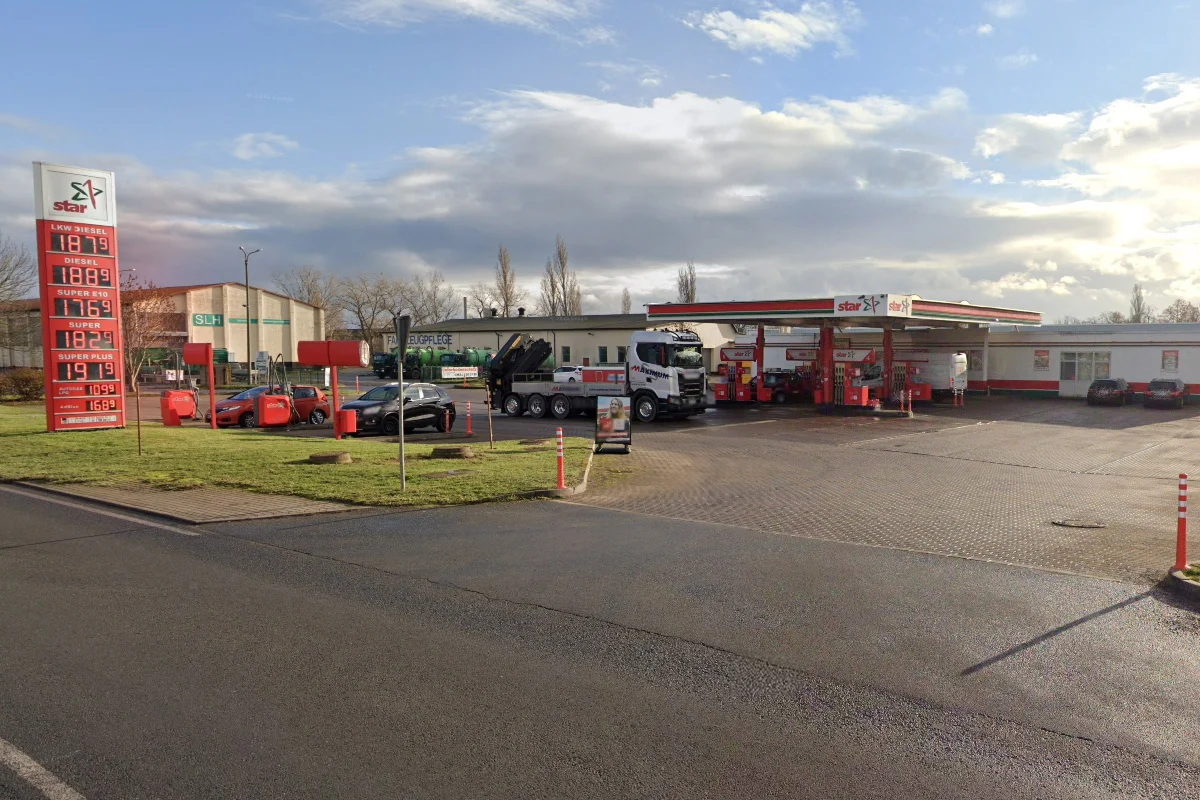 Eine Tankstelle mit einem Lkw und mehreren Autos, darunter ein rotes Fahrzeug, bei Tageslicht. Die Preisanzeige der Tankstelle ist ebenfalls sichtbar.