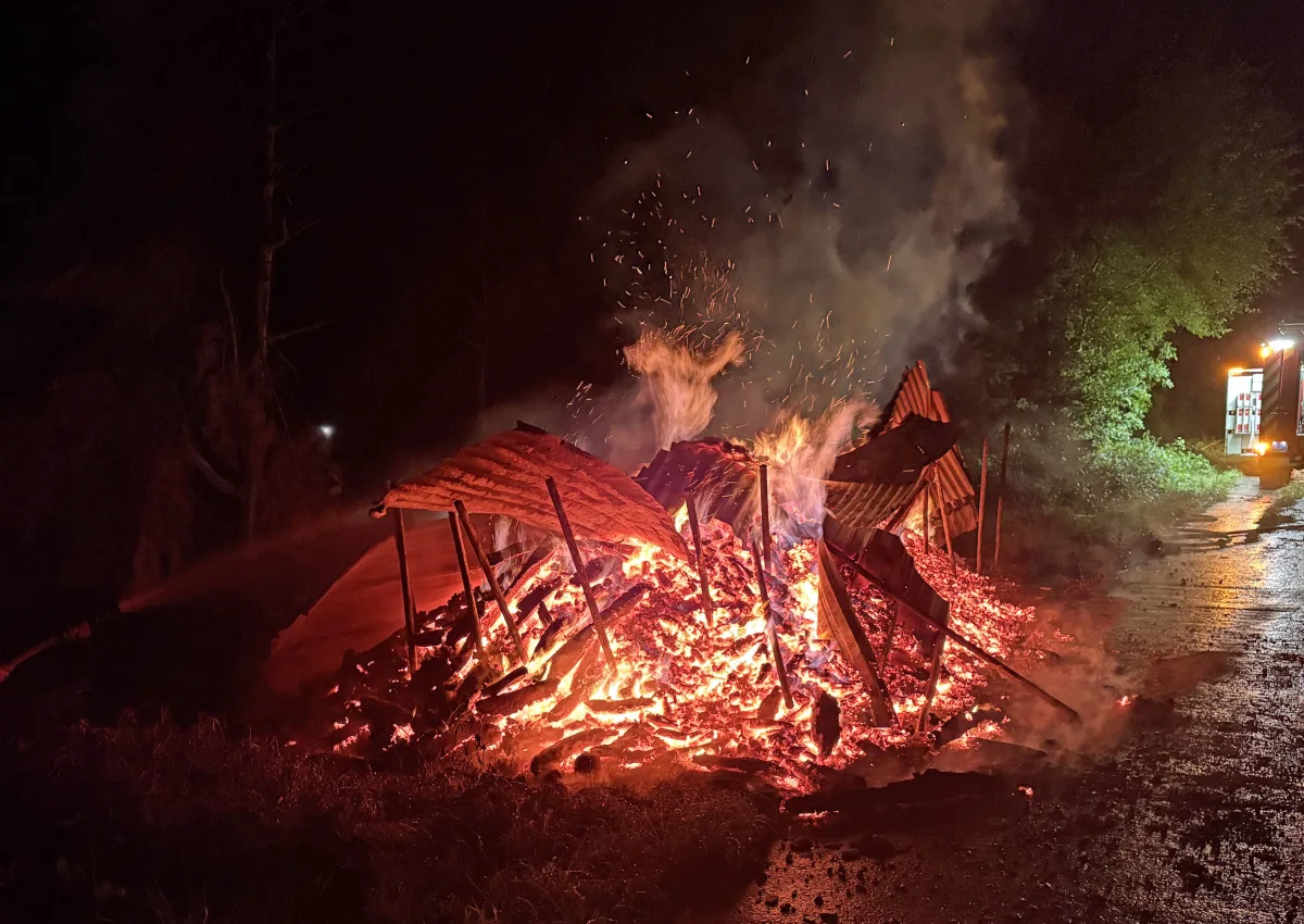 Ein intensives Feuer erfasst einen Haufen aus Holz und Blech bei Nacht. Ein Feuerwehrfahrzeug steht rechts am Bildrand auf der Straße.