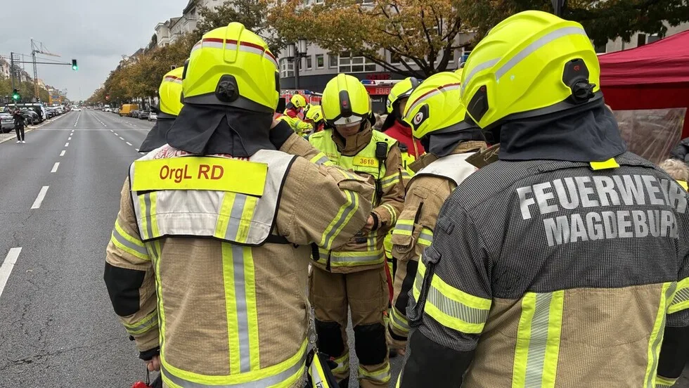 Eine Gruppe von Feuerwehrleuten in auffälligen gelben Helmen und Uniformen steht auf einer breiten Stadtstraße. Im Hintergrund sind eine Ampel und mehrere Gebäude zu sehen.