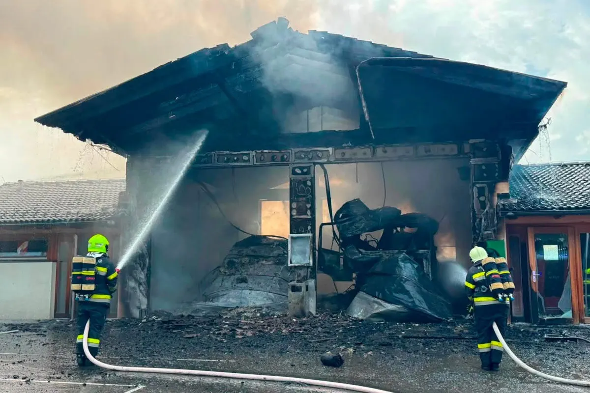 Zwei Feuerwehrleute in gelben Helmen und Atemschutzmasken spritzen Wasser auf ein brennendes, beschädigtes Gebäude. Rauch steigt auf.