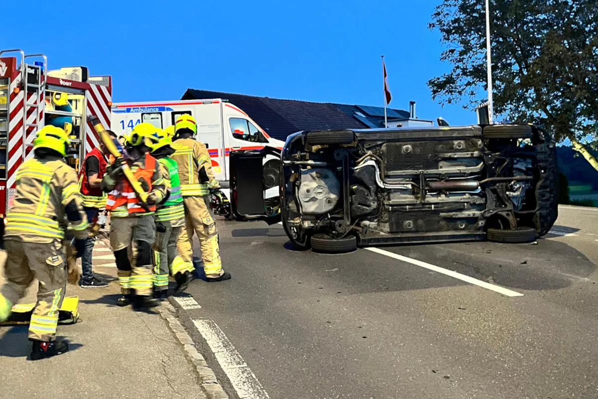 Das Bild zeigt ein auf der Seite liegendes Auto auf einer Straße, umgeben von Feuerwehrleuten in gelben Helmen. Im Hintergrund sind Einsatzfahrzeuge zu sehen.
