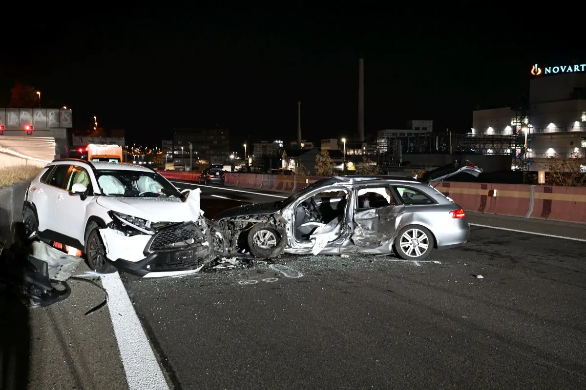 Das Bild zeigt zwei stark beschädigte Fahrzeuge nach einem Unfall auf einer nächtlichen Autobahn. Die Straße ist beleuchtet und im Hintergrund sind Gebäude zu sehen.