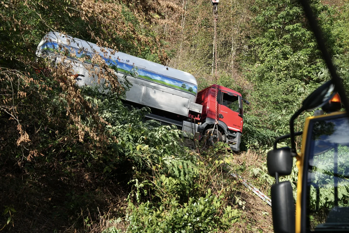Ein roter Lastwagen befindet sich in einem Waldstück und wird von einem Kran aus einer steilen Böschung gehoben. Im Vordergrund ist ein weiteres Fahrzeug zu sehen.