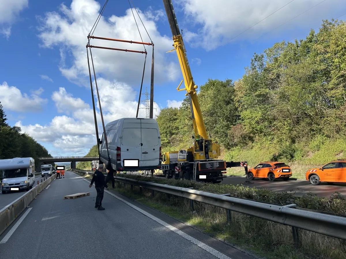 Ein gelber Kran hebt einen weißen Transporter über die Leitplanke auf einer Autobahn. Mehrere orangefarbene Autos stehen auf der anderen Fahrbahnseite.