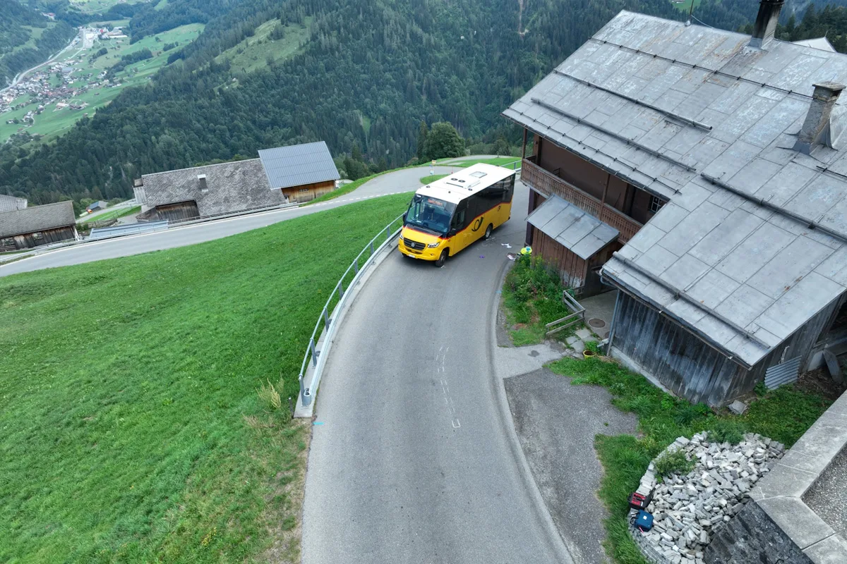 Ein gelber Postbus fährt eine enge kurvige Straße bergab neben einem alten Holzhaus in alpiner Landschaft.