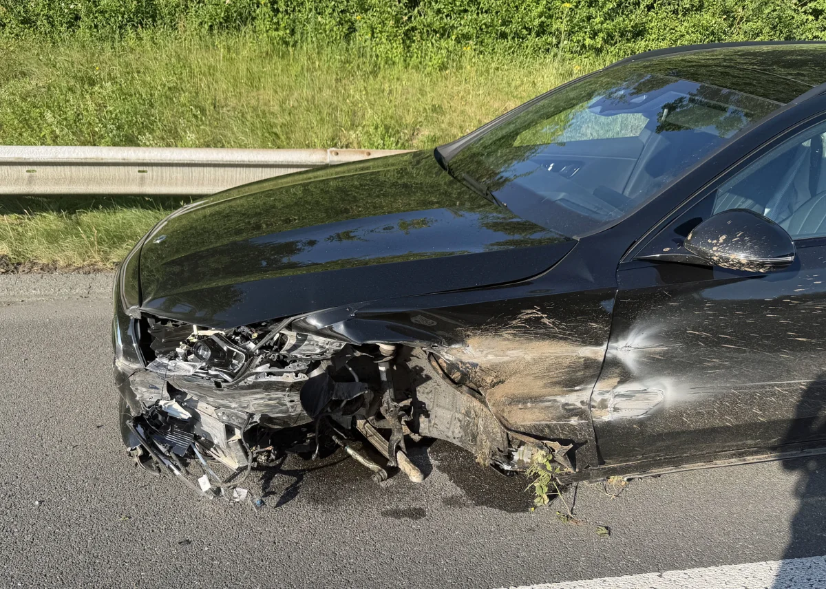 Ein schwarzes Auto mit stark beschädigter Front steht am rechten Straßenrand vor einer Leitplanke und grünem Gebüsch im Hintergrund.