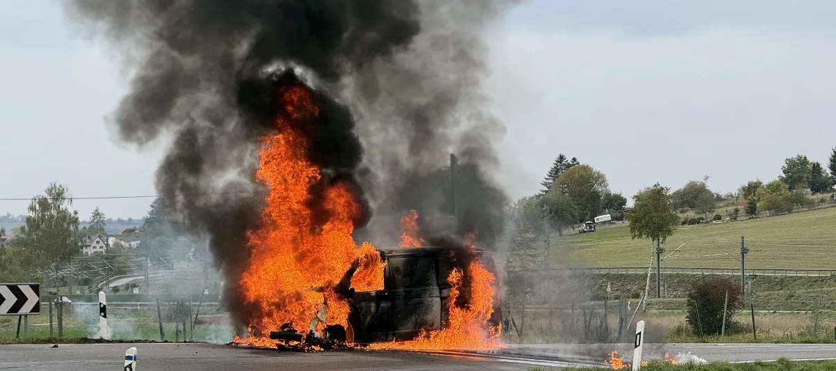 Ein Fahrzeug steht neben einer Landstraße in Flammen und erzeugt schwarzen Rauch, während die Umgebung ländlich wirkt.