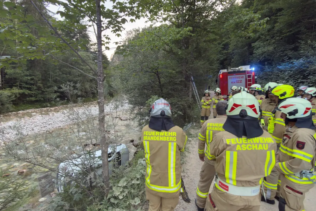 Eine Gruppe von Feuerwehrleuten in Schutzkleidung steht auf einem Pfad neben einem umgestürzten Fahrzeug in der Nähe eines Flusses. Im Hintergrund ist ein Feuerwehrfahrzeug sichtbar.