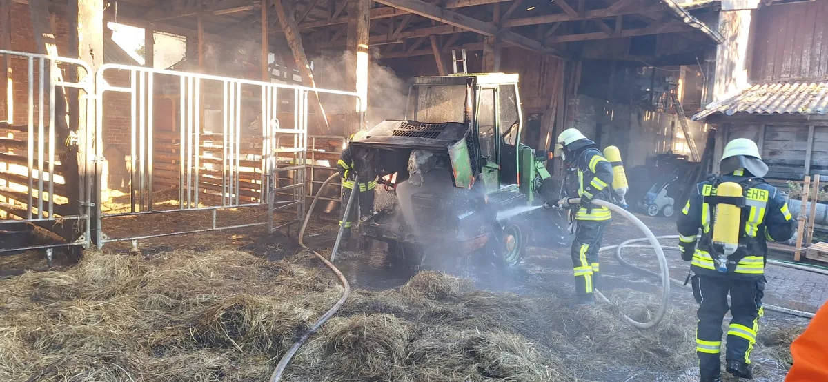 Drei Feuerwehrleute in Schutzkleidung löschen mit Schläuchen einen brennenden Traktor in einer Scheune. Rauch steigt auf und Heu ist am Boden verteilt.