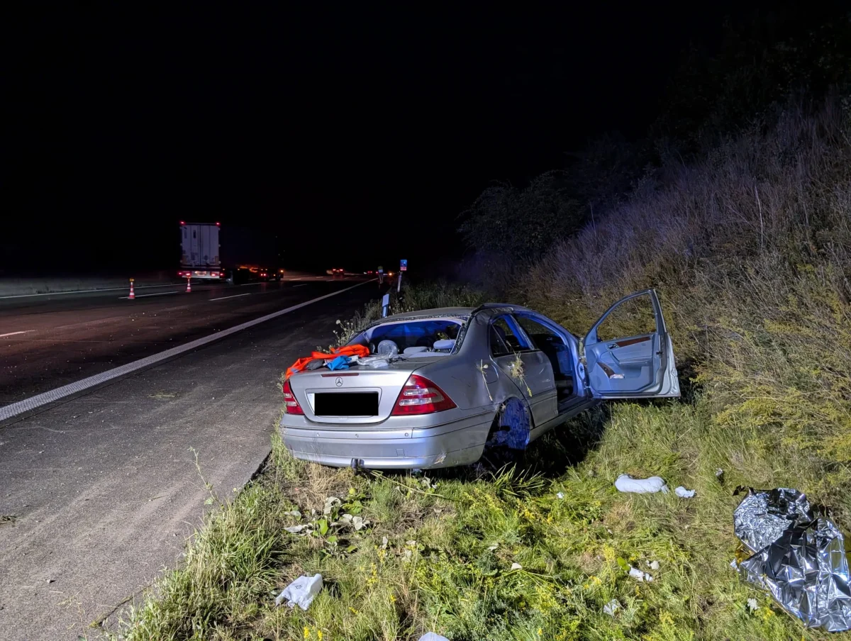 Ein silberner Mercedes steht beschädigt im Gras neben einer Autobahn. Die Fahrzeugtür ist offen und das Auto ist nach einem Unfall verlassen.