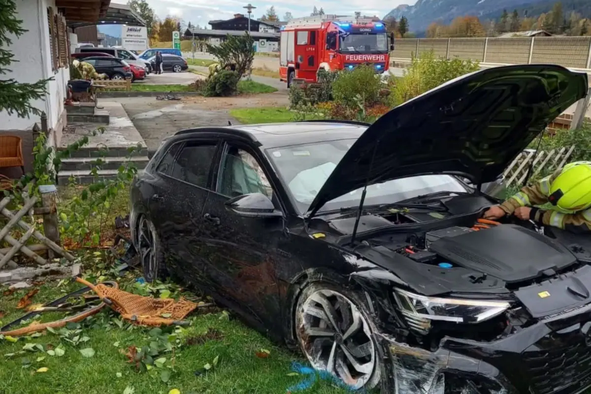 Ein schwarzes Auto steht mit geöffneter Motorhaube im Garten eines Hauses. Ein Feuerwehrmann arbeitet am Fahrzeug, im Hintergrund ein Feuerwehrauto.