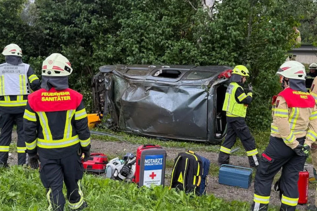 Mehrere Feuerwehrleute stehen um ein umgestürztes Auto, das am Straßenrand liegt. Rettungsausrüstung ist in der Nähe.