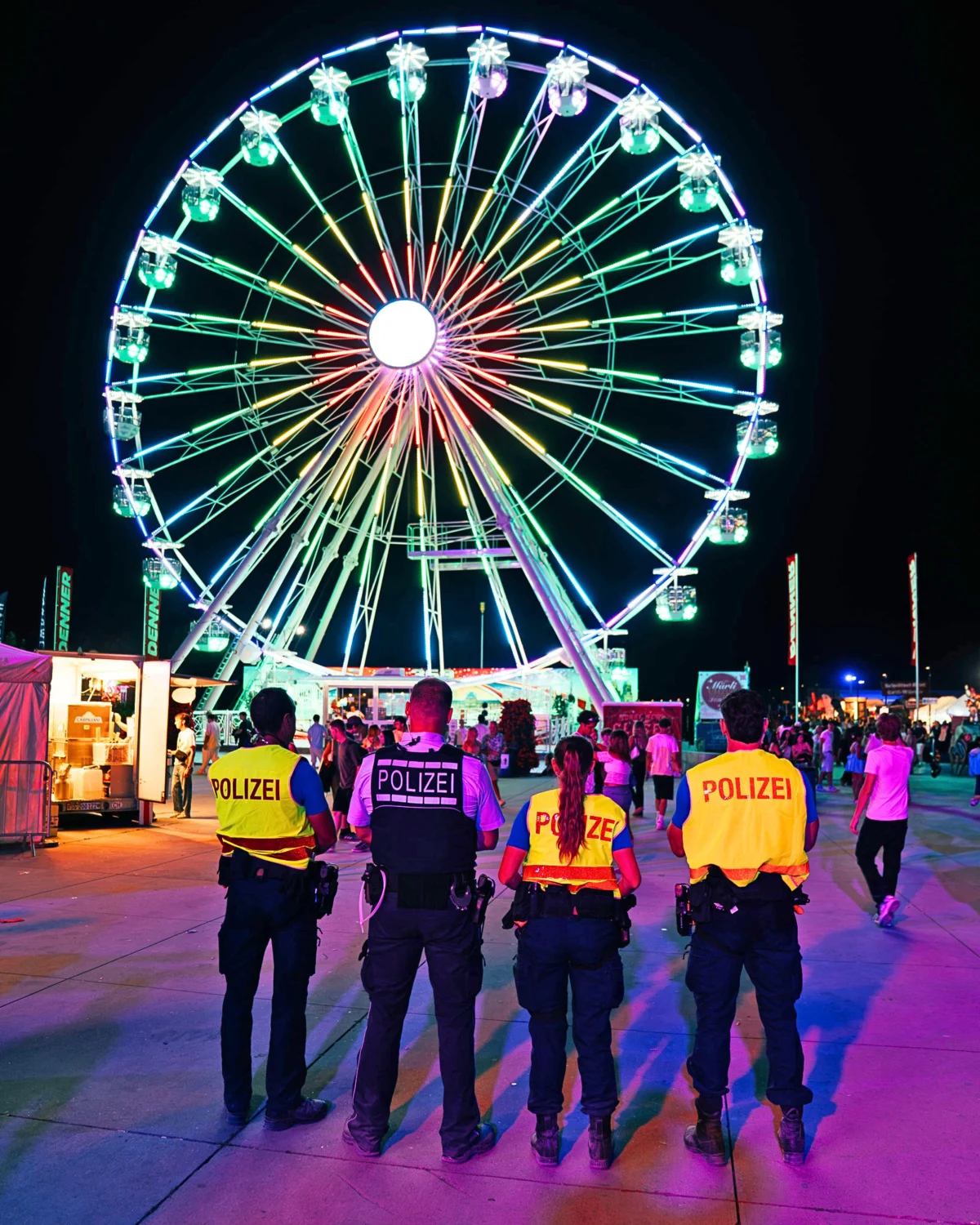 Vier Polizisten stehen bei Nacht vor einem großen, bunt beleuchteten Riesenrad. Die Umgebung wirkt wie ein belebter Rummelplatz.