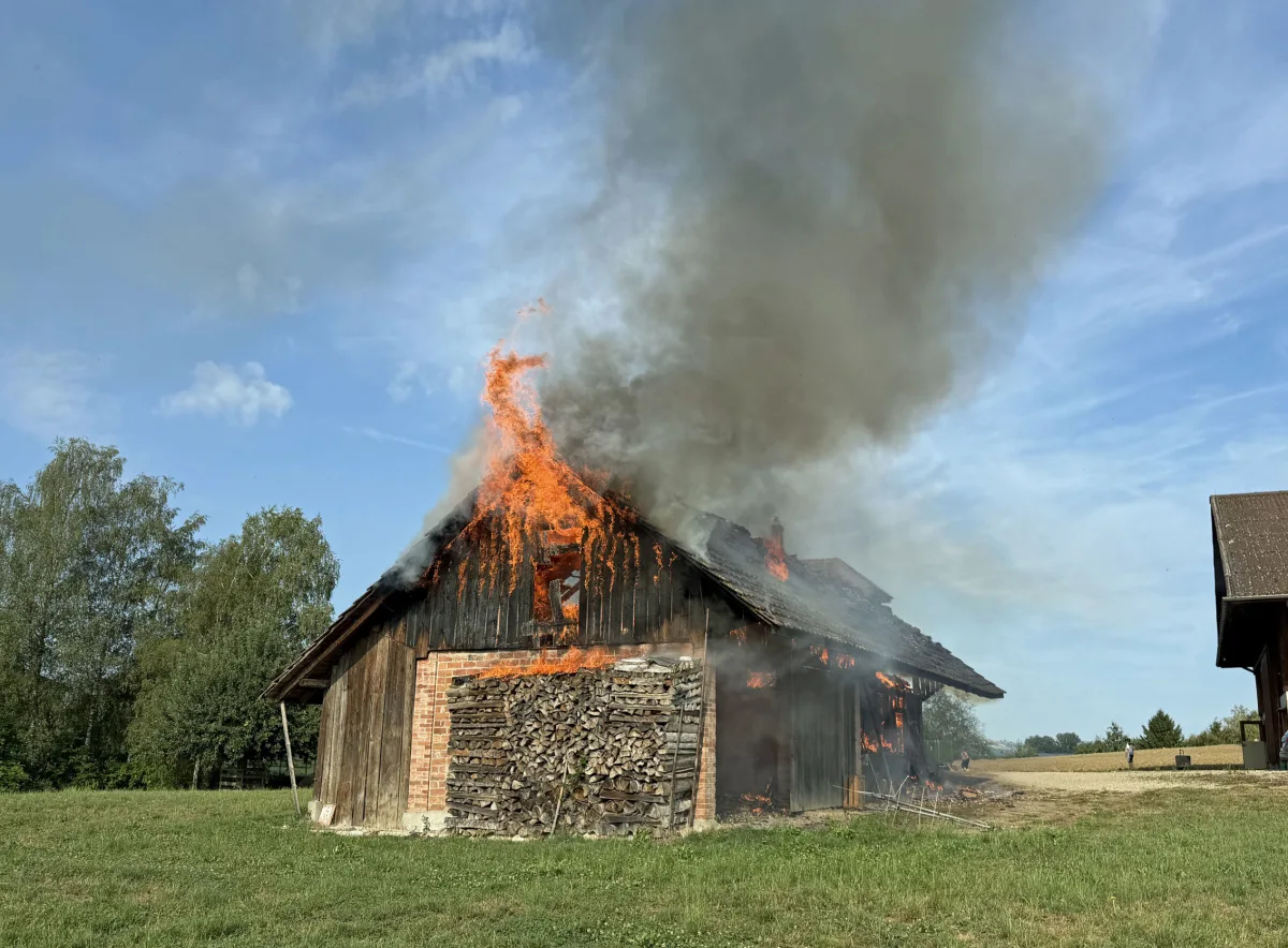 Ein ländliches Holzgebäude steht in Flammen, wobei Rauch vom Dach aufsteigt. Im Hintergrund sind Bäume und ein weiteres Gebäude sichtbar.