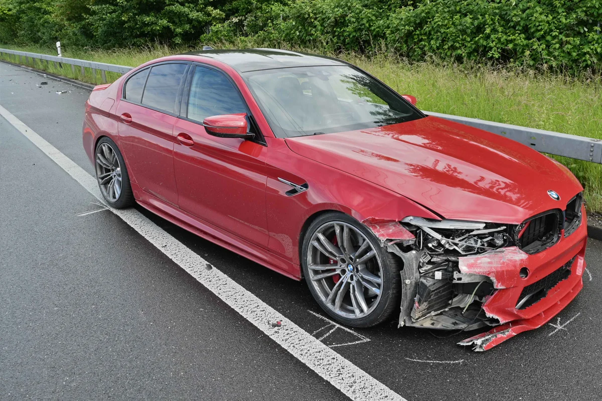 Ein roter BMW steht auf einer Autobahn mit erheblichem Frontschaden. Der Wagen befindet sich in der Nähe einer Leitplanke und die Straße ist asphaltiert.