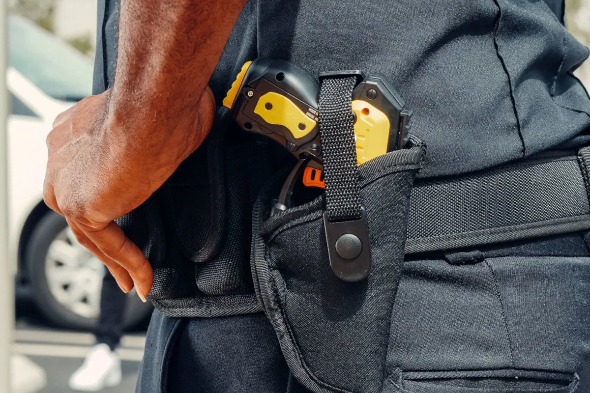 Ein Polizist trägt ein Elektroschockgerät in einem Holster an seiner Hüfte, nahe zu einem parkenden Auto.