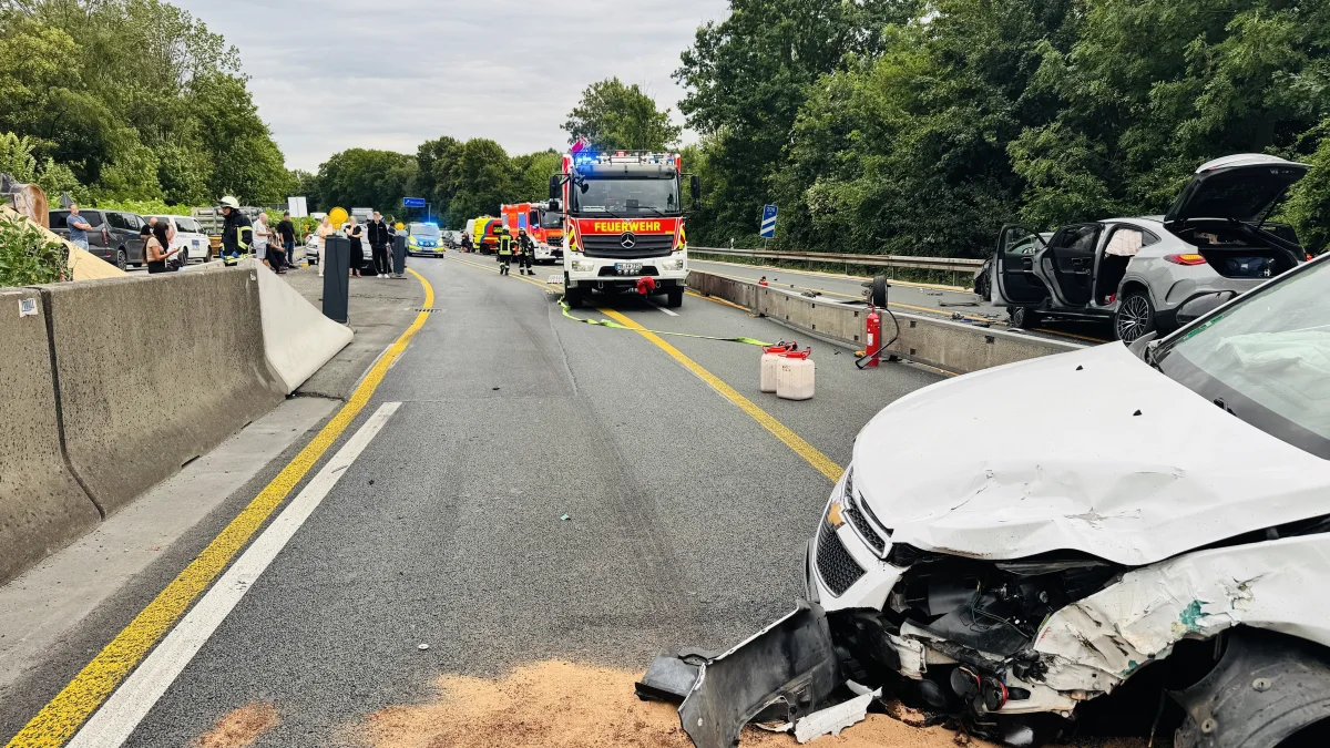 An einer Autobahn sind ein beschädigtes Auto, ein Feuerwehrfahrzeug und mehrere Einsatzkräfte zu sehen. Der Unfall hat zur Sperrung der mittleren Fahrspur geführt.
