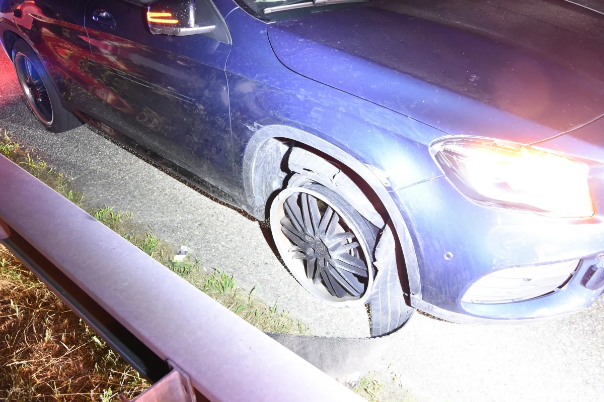 Ein blaues Auto steht auf einer Straße bei Nacht. Das Vorderrad ist stark beschädigt, und das Fahrzeug steht neben einer Leitplanke.