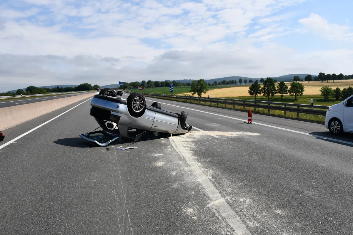 Ein silbernes Auto liegt auf dem Dach auf einer Autobahn mit Verkehrskegeln und einem weißen Fahrzeug in der Nähe. Im Hintergrund sind Felder und Bäume sichtbar.