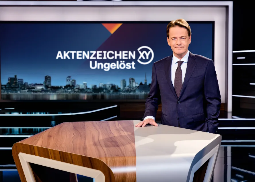 Ein Moderator steht in einem schicken Anzug neben einem modernen Studiotisch, im Hintergrund eine nächtliche Skyline auf einem Bildschirm.