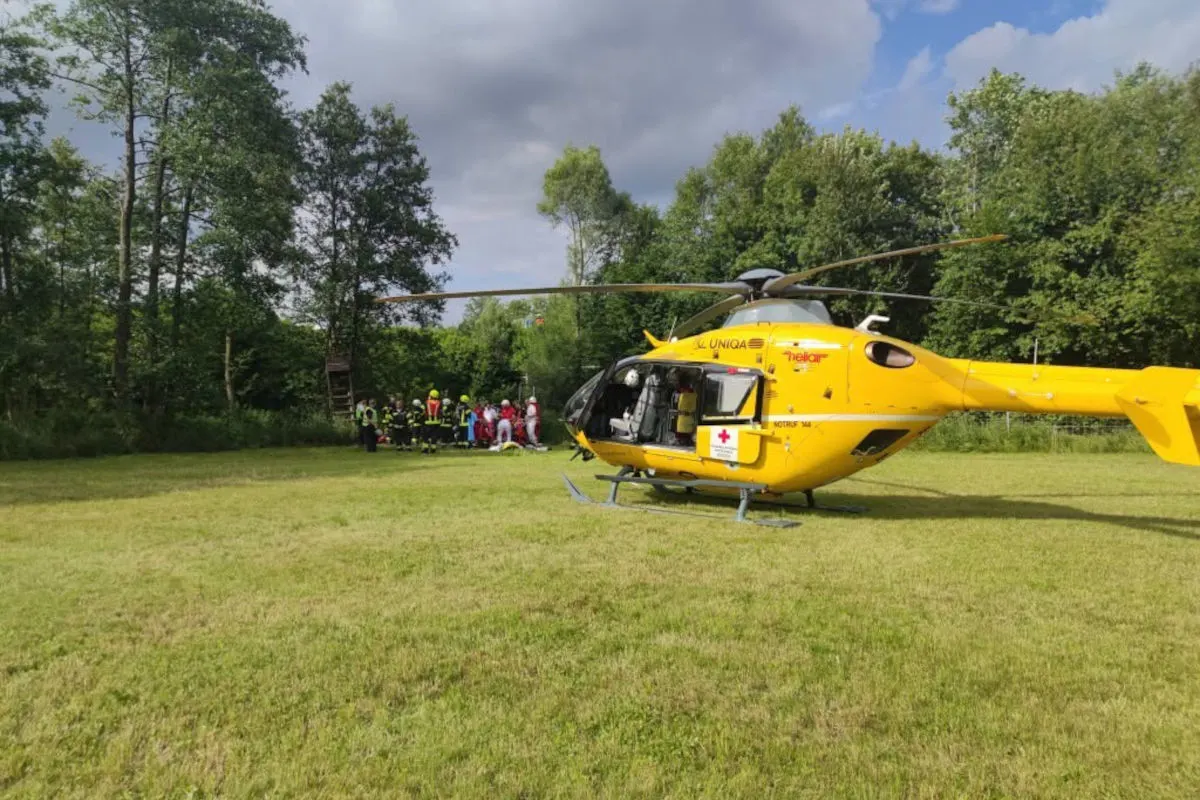 Ein gelber Rettungshubschrauber steht auf einer Wiese, während mehrere Einsatzkräfte in Schutzkleidung am Bildrand zu sehen sind. Der Hintergrund ist von Bäumen gesäumt, unter einem sonnigen Himmel mit einigen Wolken.