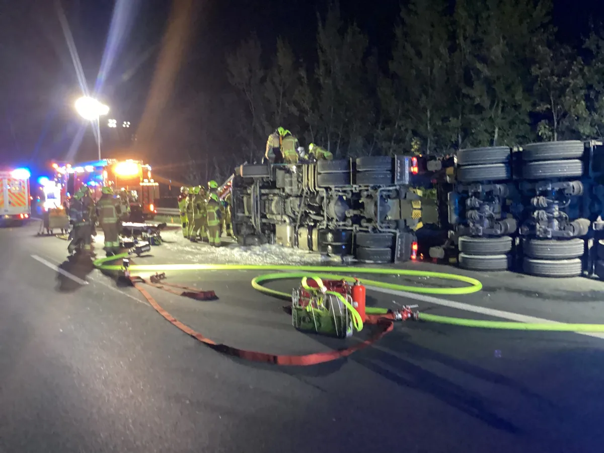 Ein umgekippter Lkw liegt quer über die Straße. Feuerwehrleute sind vor Ort, beleuchtet von Einsatzfahrzeugen.
