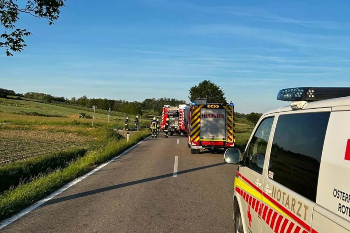 Mehrere Feuerwehrfahrzeuge und ein Notarztfahrzeug stehen auf einer geraden Landstraße. Die Umgebung ist ländlich mit Feldern und Bäumen, der Himmel ist klar und blau.