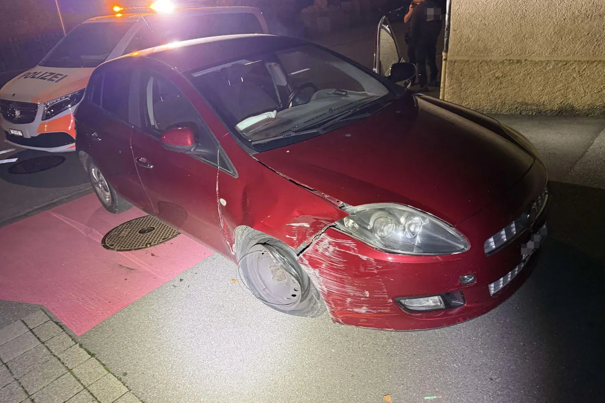 Ein rotes Auto zeigt deutliche Kratzer und Dellen an der Vorderseite. Im Hintergrund steht ein Polizeiwagen mit leuchtendem Licht.