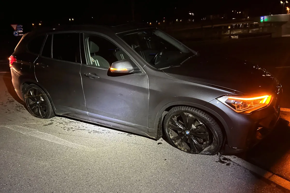 Ein dunkelgrauer BMW steht nachts auf einer Straße, der rechte Vorderreifen ist platt. Das Fahrzeug hat die Blinker eingeschaltet.