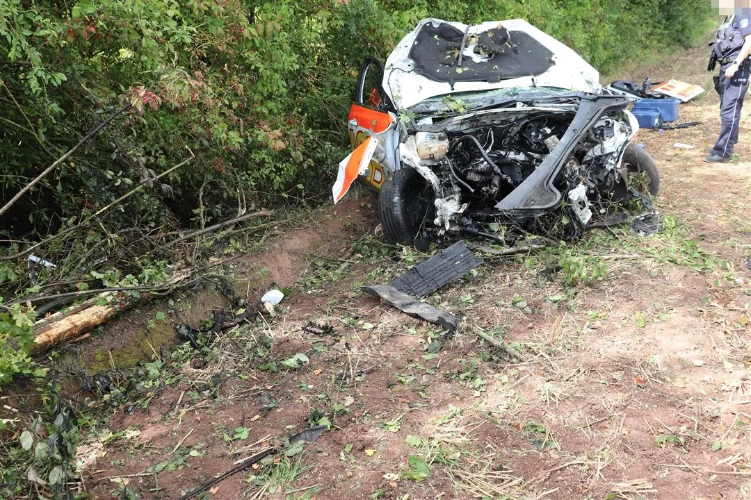 Das Bild zeigt ein stark beschädigtes Fahrzeug im Vordergrund, das von der Fahrbahn abgekommen ist und am Straßenrand im Grünen liegt. Ein Trümmerfeld und Pflanzen sind sichtbar, was auf einen heftigen Aufprall schließen lässt.