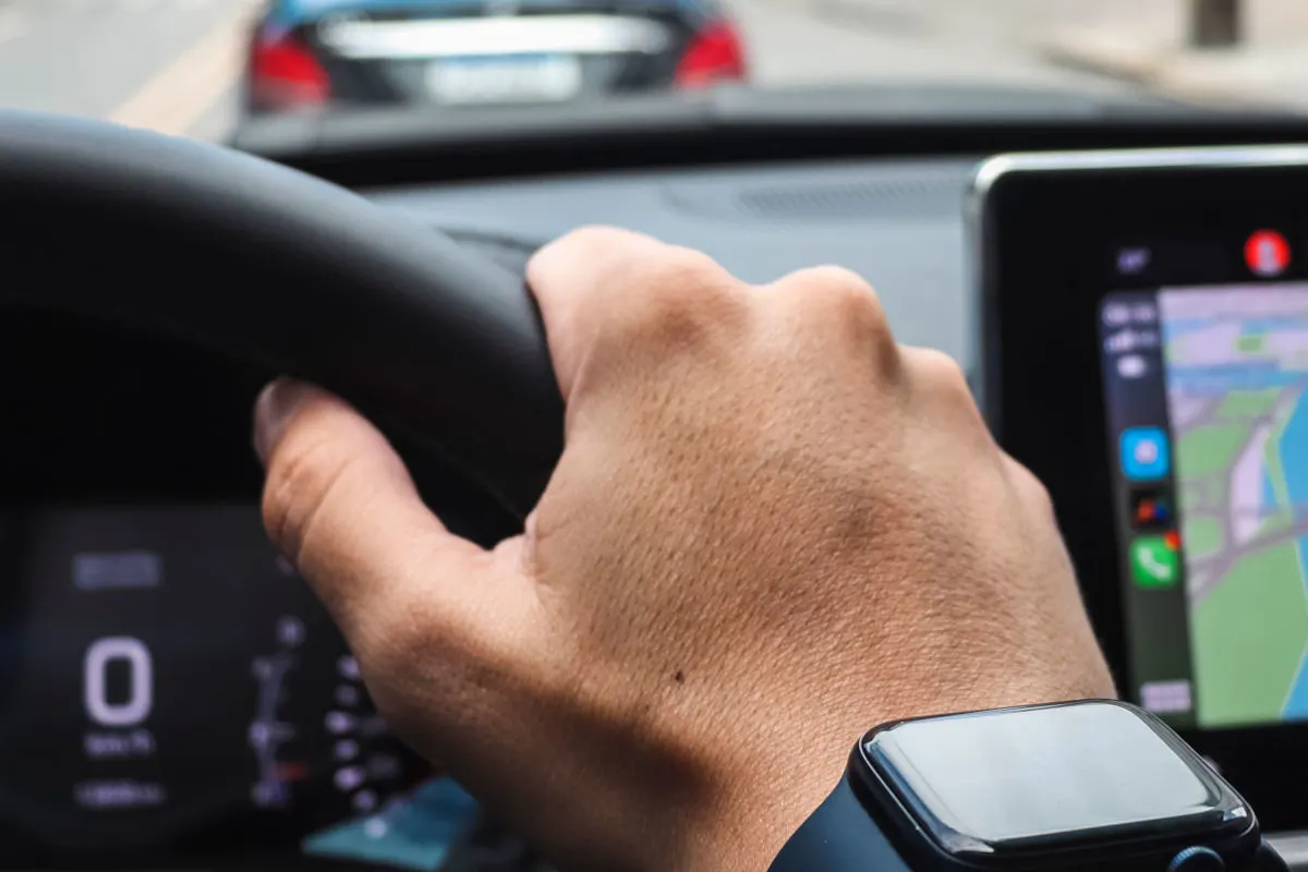 Eine Hand hält das Lenkrad eines Autos, während ein Bildschirm mit Navigationssystem im Hintergrund sichtbar ist. Ein weiteres Fahrzeug ist voraus auf der Straße zu erkennen.