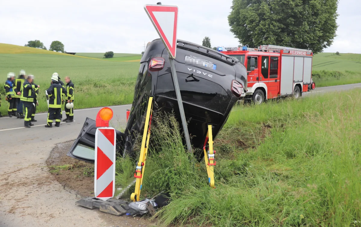 Ein Auto liegt auf der Seite neben einem Verkehrsschild auf einer Landstraße. Feuerwehrkräfte stehen im Hintergrund, während ein rotes Feuerwehrfahrzeug sichtbar ist.