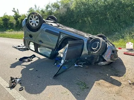 Ein Auto liegt auf dem Dach auf einer Landstraße, umgeben von verstreuten Fahrzeugteilen. Die Straße ist von Bäumen gesäumt.