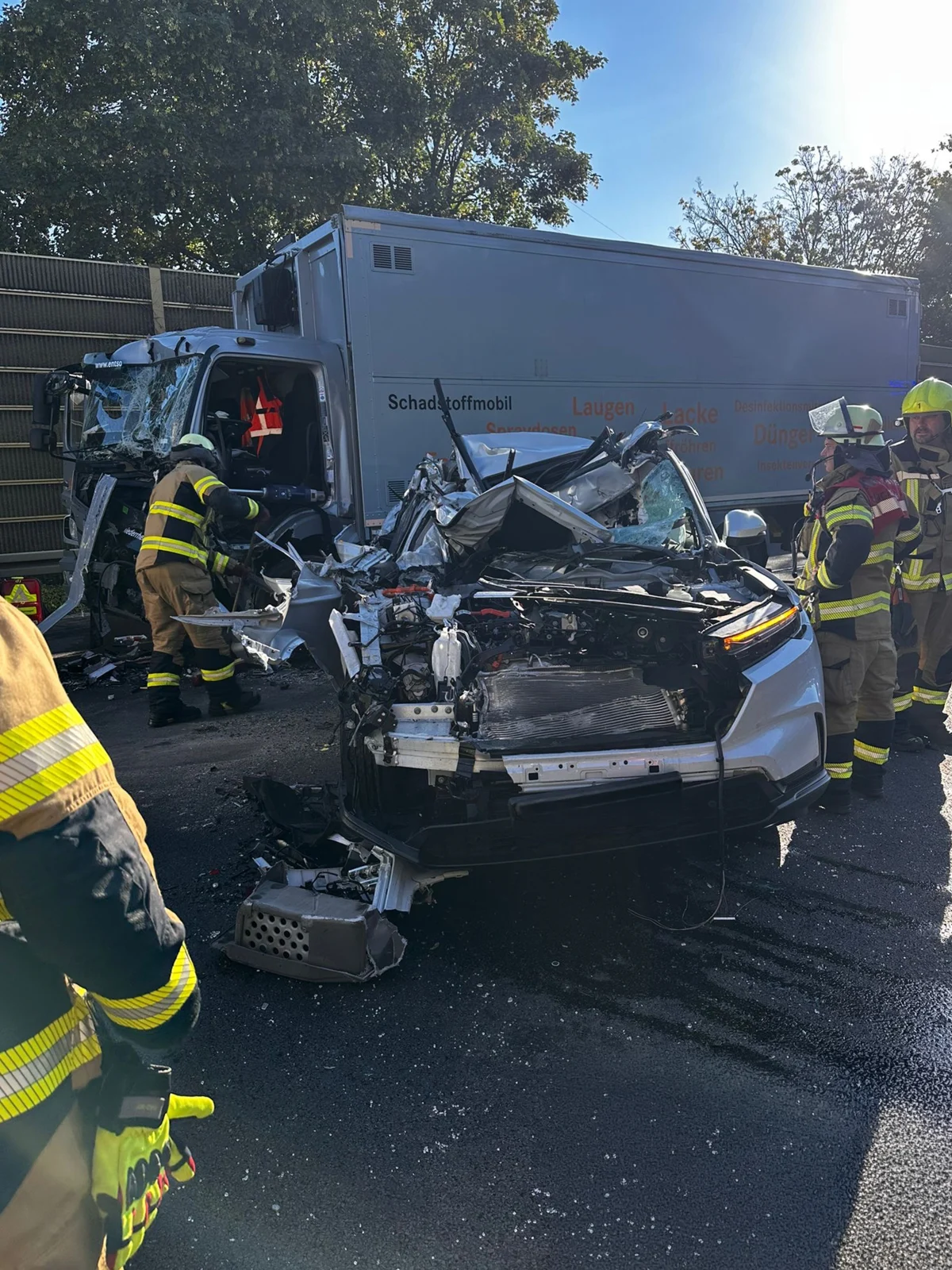 Ein stark beschädigtes Auto steht vor einem Lastwagen mit der Aufschrift 'Schadstoffmobil'. Mehrere Feuerwehrleute arbeiten an der Unfallstelle.