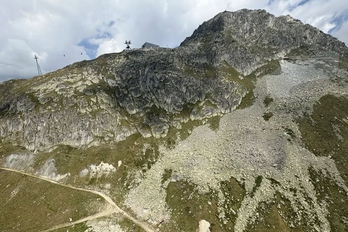 Eine felsige Berglandschaft mit einer Seilbahn ist bei teilweise bewölktem Himmel zu sehen, darunter ein schmaler Wanderpfad.