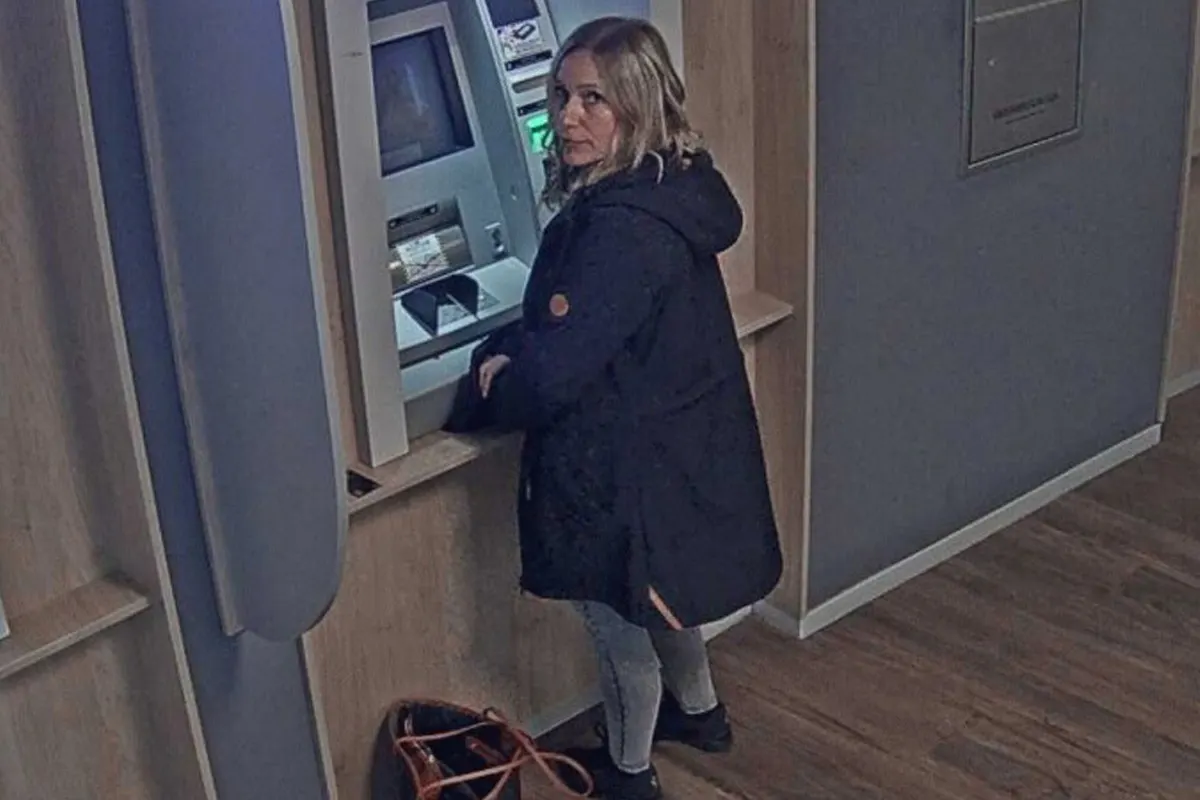 Eine blonde Frau mit einem schwarzen Mantel steht in einer Bank und bedient einen Geldautomaten. Eine Handtasche steht auf dem Boden.