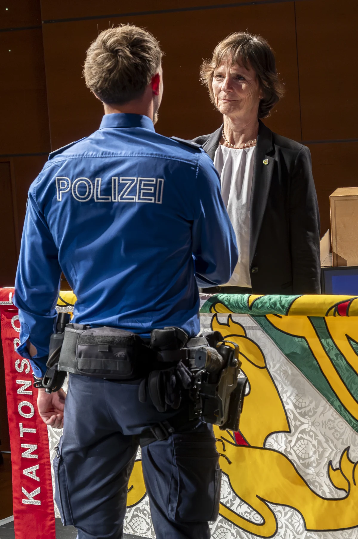 Ein Polizist in blauer Uniform, Rückenansicht, unterhält sich mit einer Frau. Im Hintergrund ist ein Wappen zu sehen.