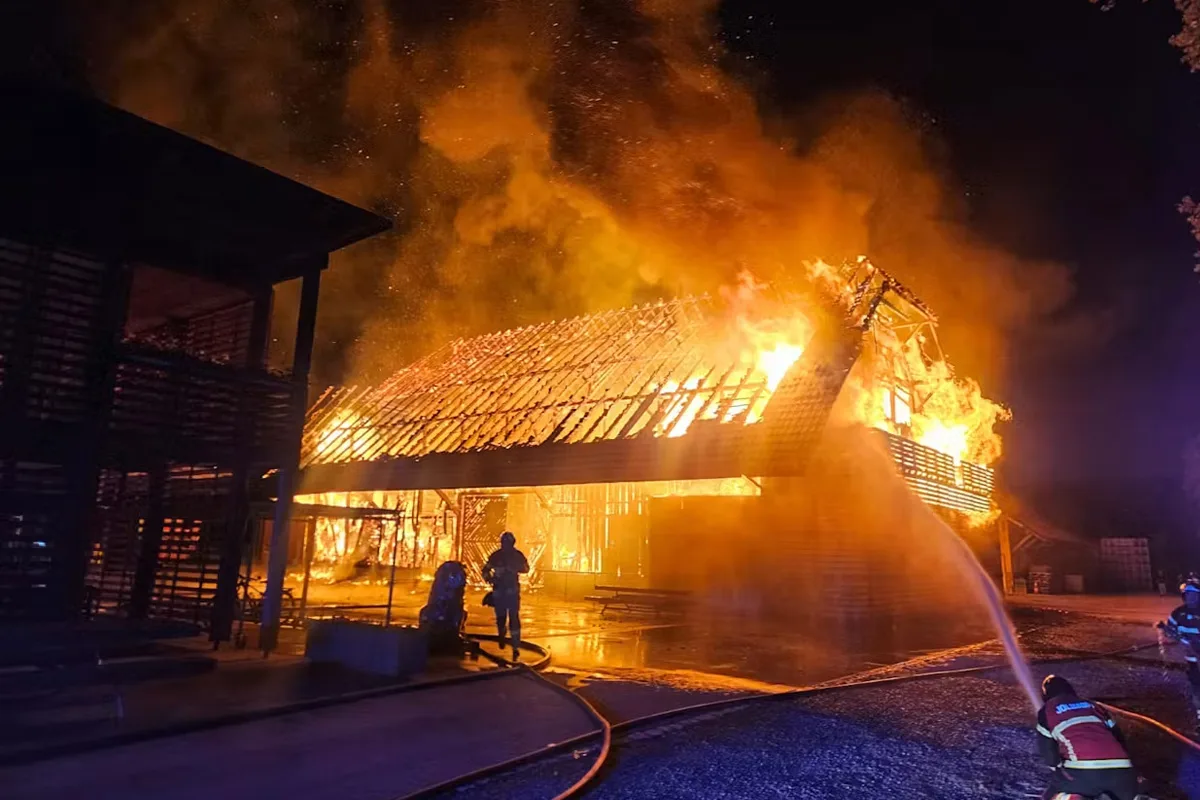Ein großes Feuer erfasst ein Gebäude, während mehrere Feuerwehrleute versuchen, die Flammen mit Schläuchen zu löschen. Der Himmel ist dunkel und die Szene wird von den Flammen beleuchtet.