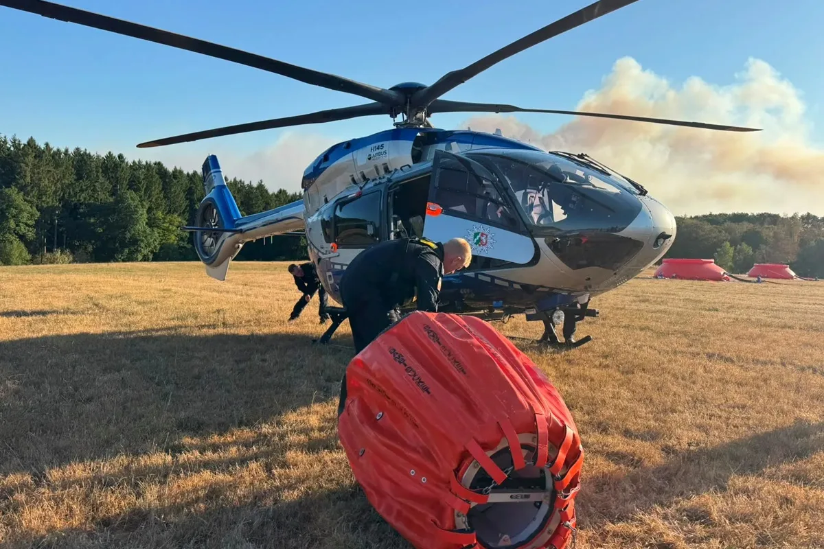 Ein Polizeihubschrauber steht auf einer Wiese. Ein Wasserbehälter liegt im Vordergrund, Personen sind neben dem Hubschrauber beschäftigt.