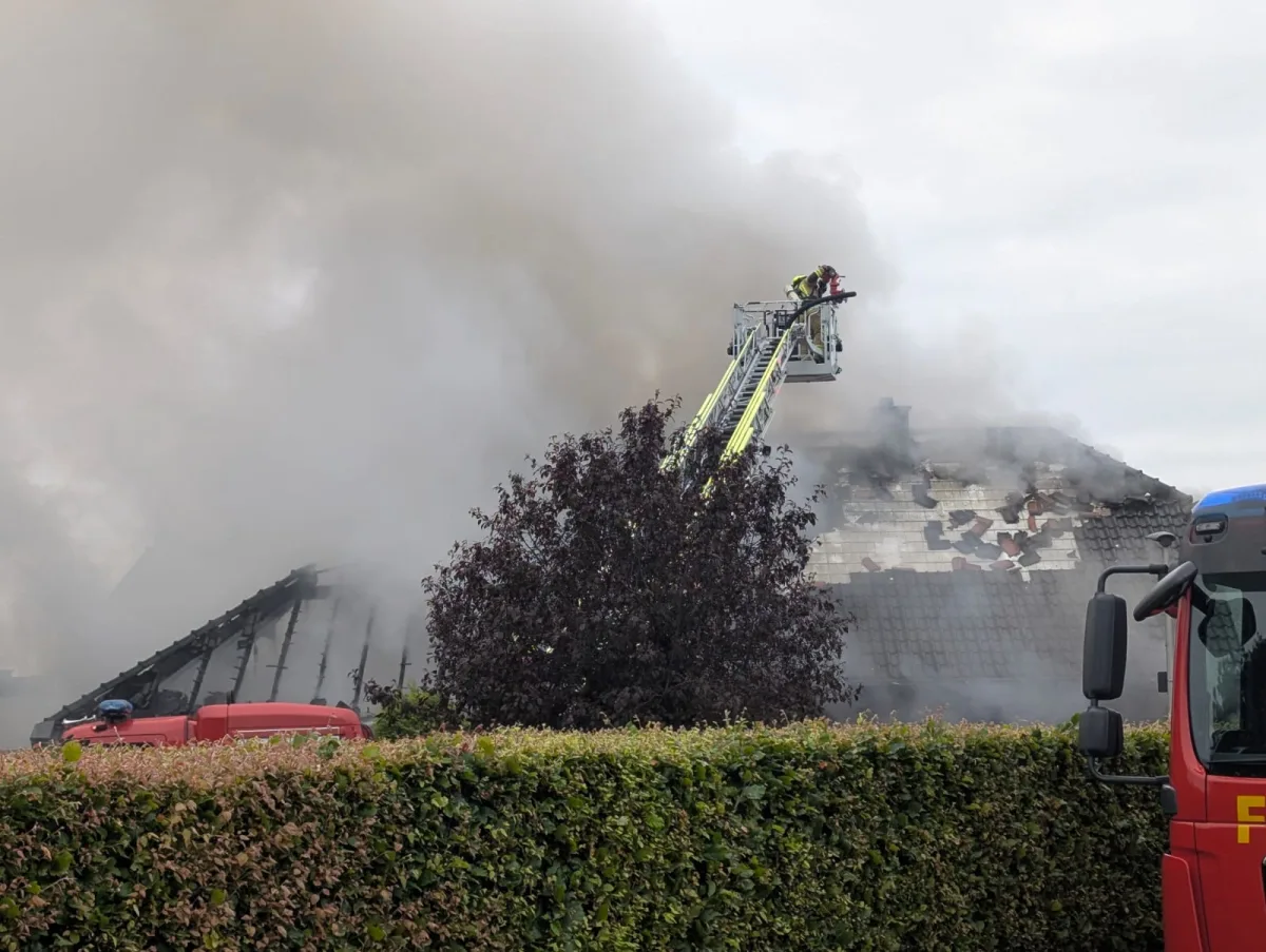 Feuerwehrleute stehen auf einer Leiter und löschen ein Feuer auf einem stark beschädigten Hausdach. Eine große Rauchentwicklung ist sichtbar.